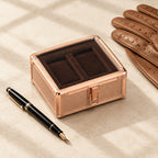 2-slot rose-gold aluminum watch box closed, beside a fountain pen and leather gloves