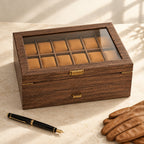 24-slot walnut wood-grain watch box closed, beside a fountain pen and leather gloves