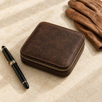 Dark brown leather watch travel case zipped shut, beside a fountain pen and leather gloves