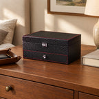 Two-tier carbon-pattern watch box on a wooden chest of drawers, beside a leather tray and a book, for scale